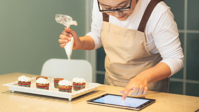 Chinese woman is decorates cupcakes and checks the recipe on iPad.