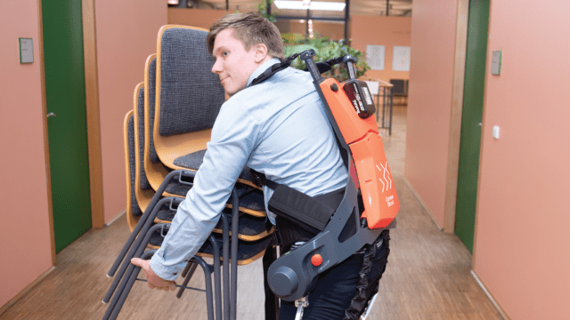 Photograph shows a man demonstrating how a robotic exoskeleton could help workers lift heavy loads