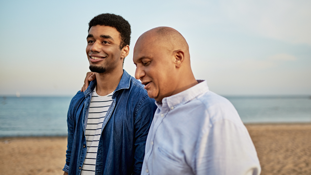 Candid outdoor portrait of mixed race man in late 40s with arm around shoulder of smiling son in early 20s as they walk and talk along Barceloneta Beach. Credit xavierarnau/Getty Images