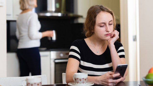 A young adult seated at a table in a home looks at her phone while an adult woman stands in the background iStock