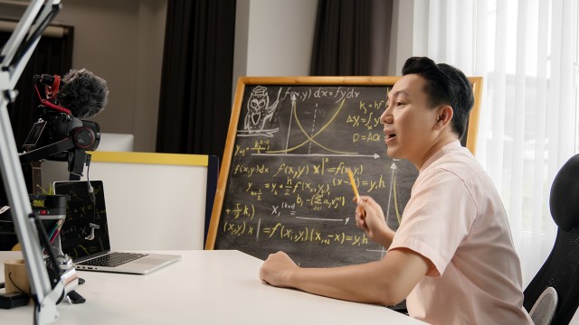 A person sitting at a desk speaks into a camera and points to a chalkboard with equations on it. (Sarote Pruksachat via Getty Images)