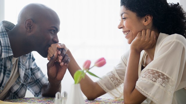 A man kisses the hand of a woman as she laughs. Both are seated at a table that has a vase of tulips. (Getty Images)