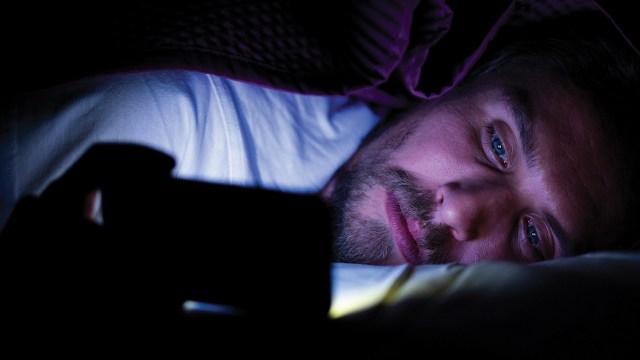 A person lying under the covers in a dark room and looking at a smartphone. (Cavan Images via Getty Images)