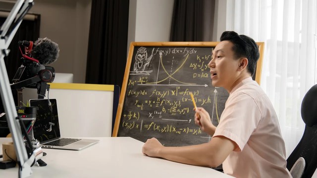 A person sitting at a desk speaks into a camera and points to a chalkboard with equations on it. (Sarote Pruksachat via Getty Images)