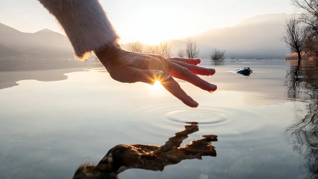 Detail of hand touching and caressing water surface of beautiful lake at sunset, mountain view.