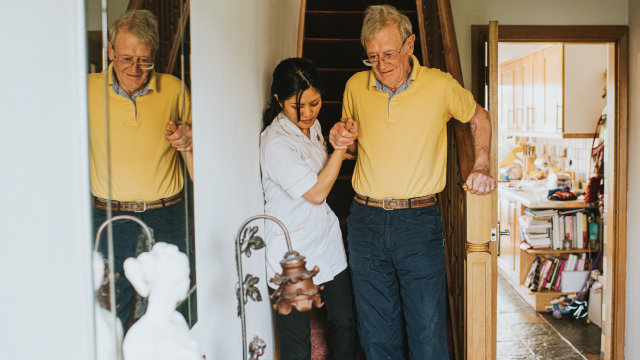 A young female care assistant helps an elderly man, in his 80s, down a narrow staircase in his own home.